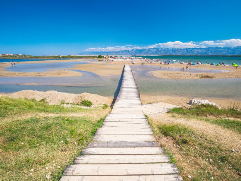 Beim Strand von Kraljicina Plaza führt ein Steg durch seichtes Wasser an den feinen Strand.