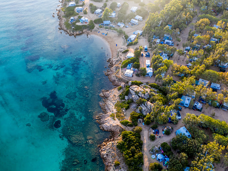Drohnenansicht des Campingplatzes Baia Holiday Isuledda mit kristallklarem Wasser vor der Küste mit Campingfahrzeugen