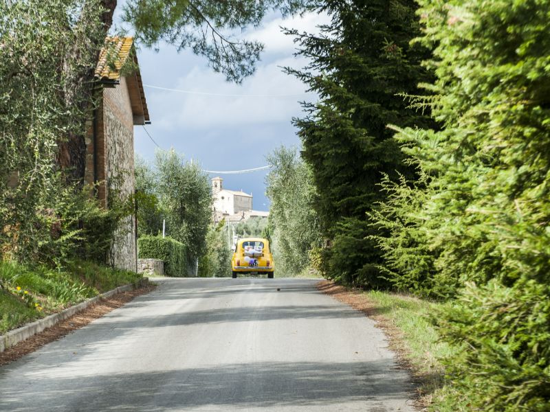 kleines gelbes Auto auf schmaler Straße von Bäumen gesäumt und im Hintergrund typisch italienische Backstein-Gebäude