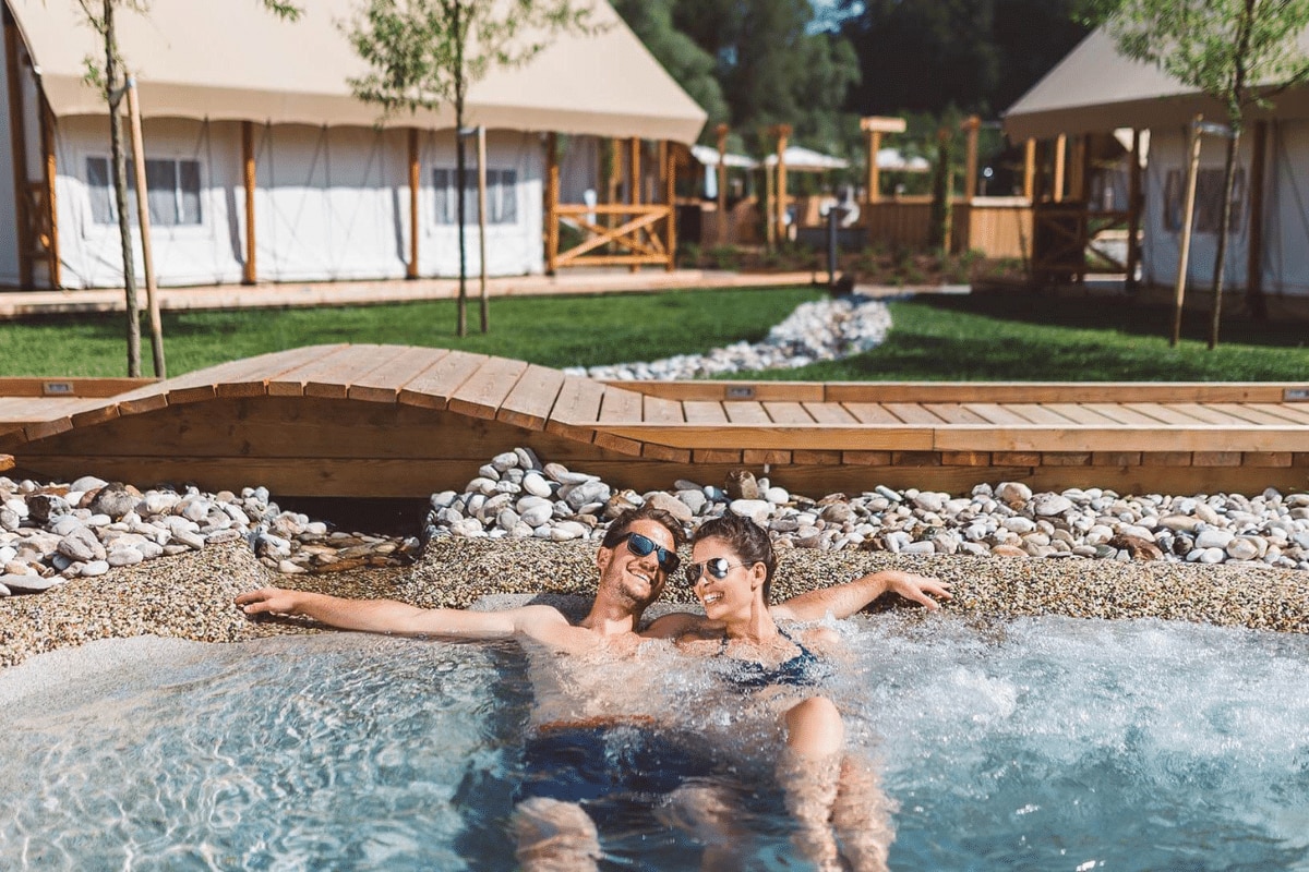 Camping Terme Olimia Natura – Slovenien (Podčetrtek) Paar liegt im Whirlpool, im Hintergrund Mietunterkünfte